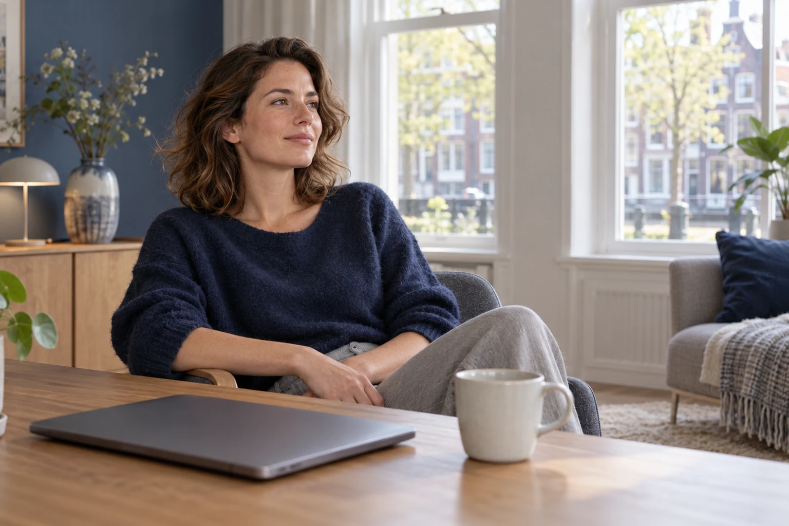 Vrouw ontspannen in stoel bij raam, mok en gesloten laptop op tafel nearby
